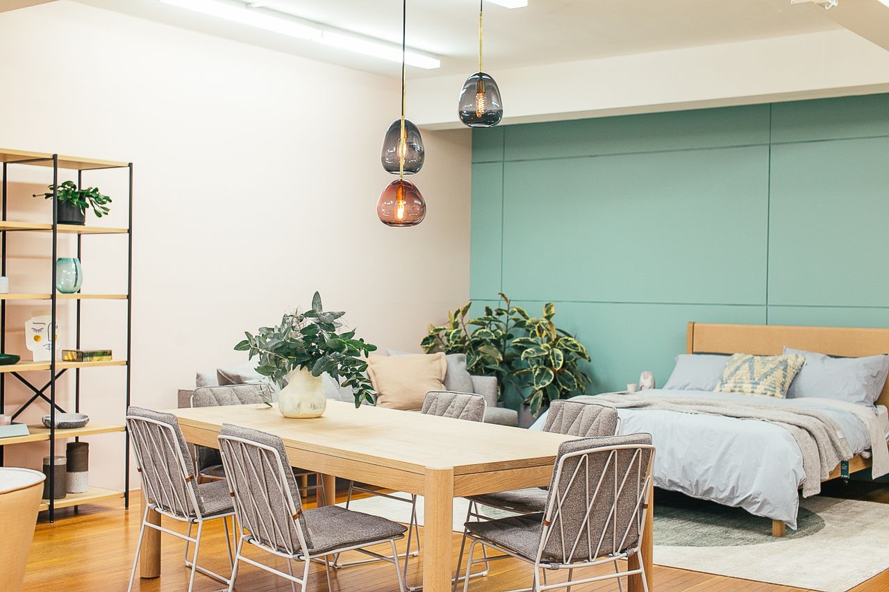 services-01 Modern light studio decorated with wooden table with vase with plant and chairs under lamps near couch and bed with carpet near shelves